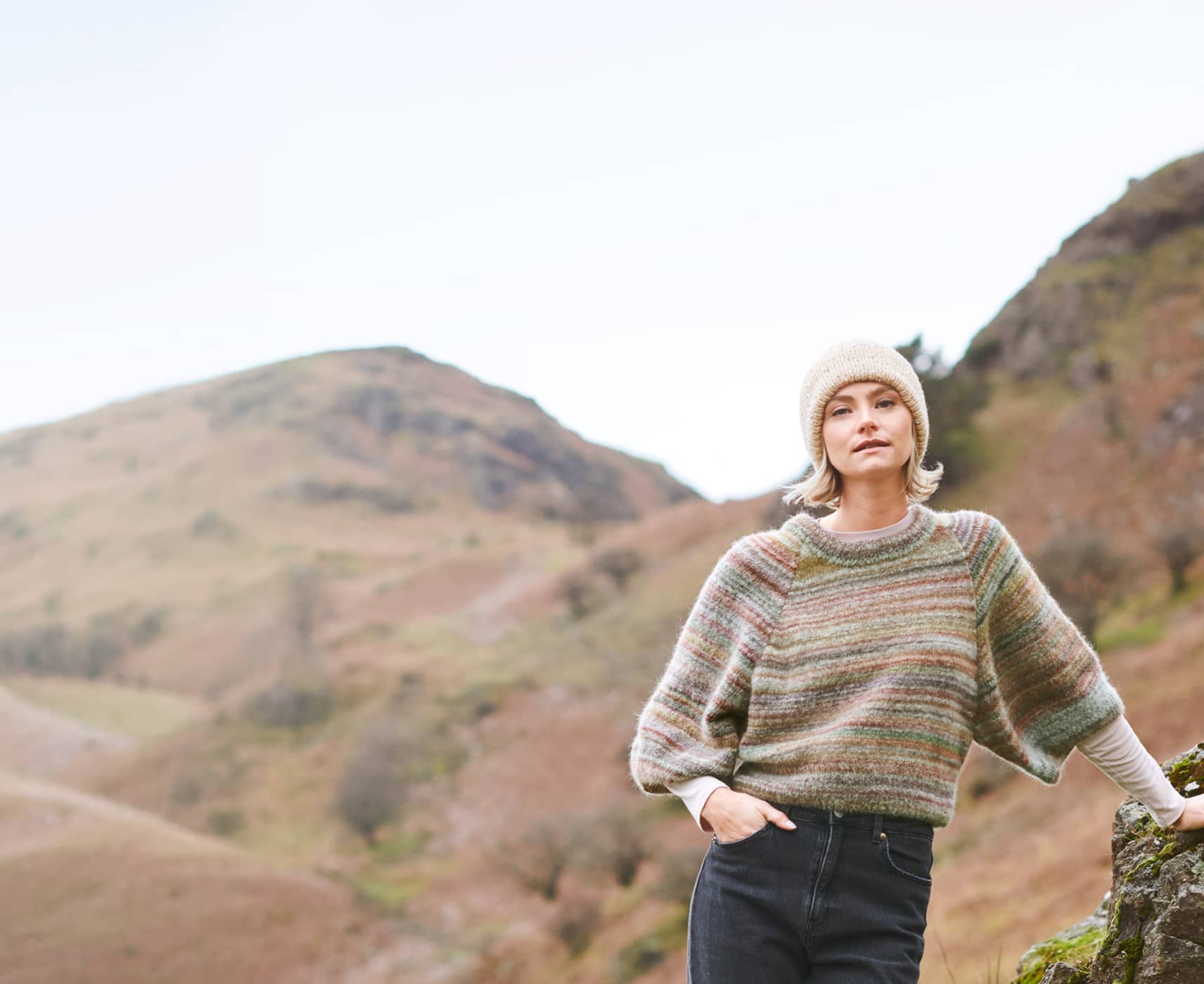 Eine Frau trägt ein herbstliches Outfit mit gestricktem Pullover und Mütze und steht vor einer Landschaft mit Bergen.