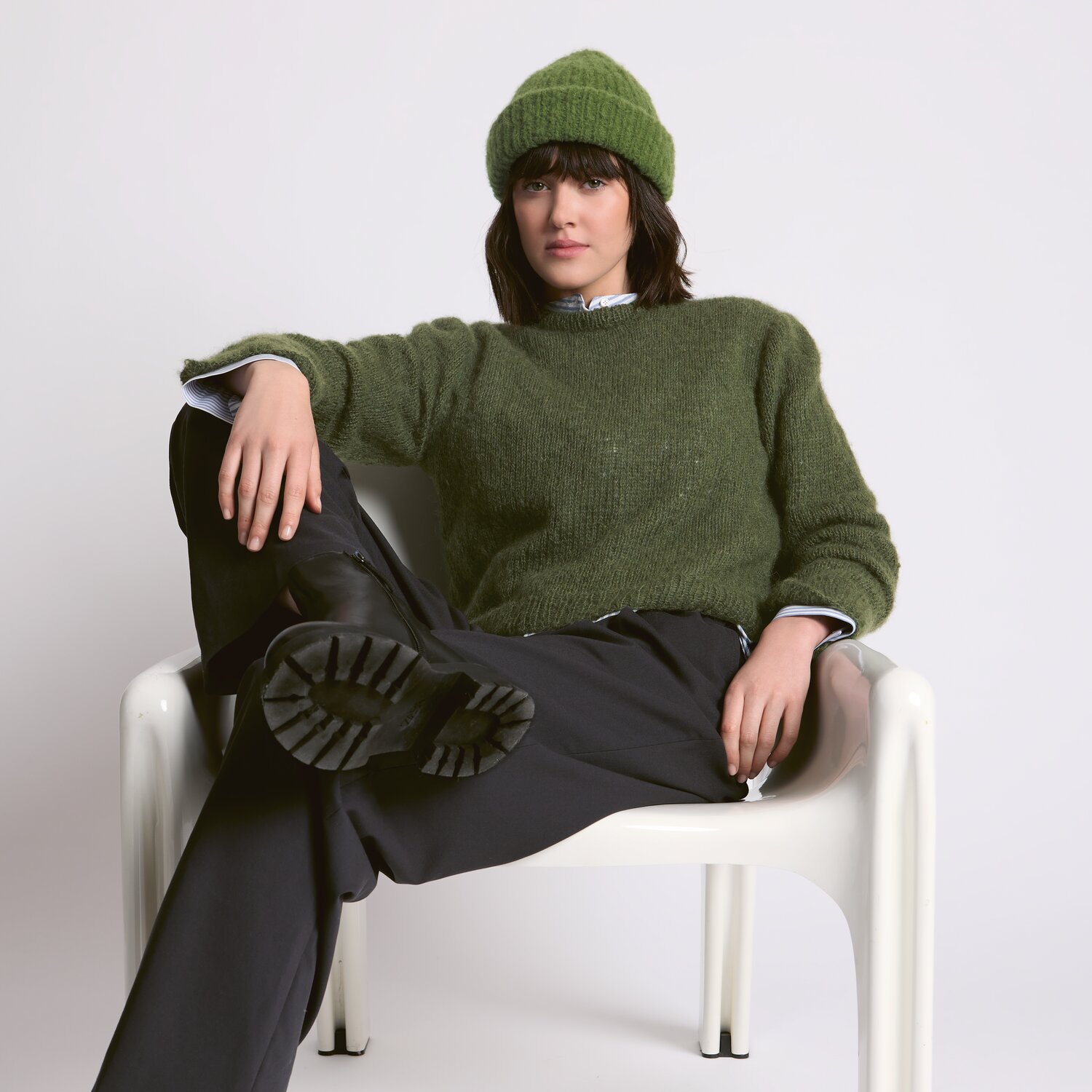 Strickset Pullover Modell 21 aus Luxury Knits - Premium Natural Fibres von Rico Design Strickset Pullover Modell 21 aus Luxury Knits: Frau in olivgrünem Pullover und Mütze sitzt lässig auf weißem Stuhl.