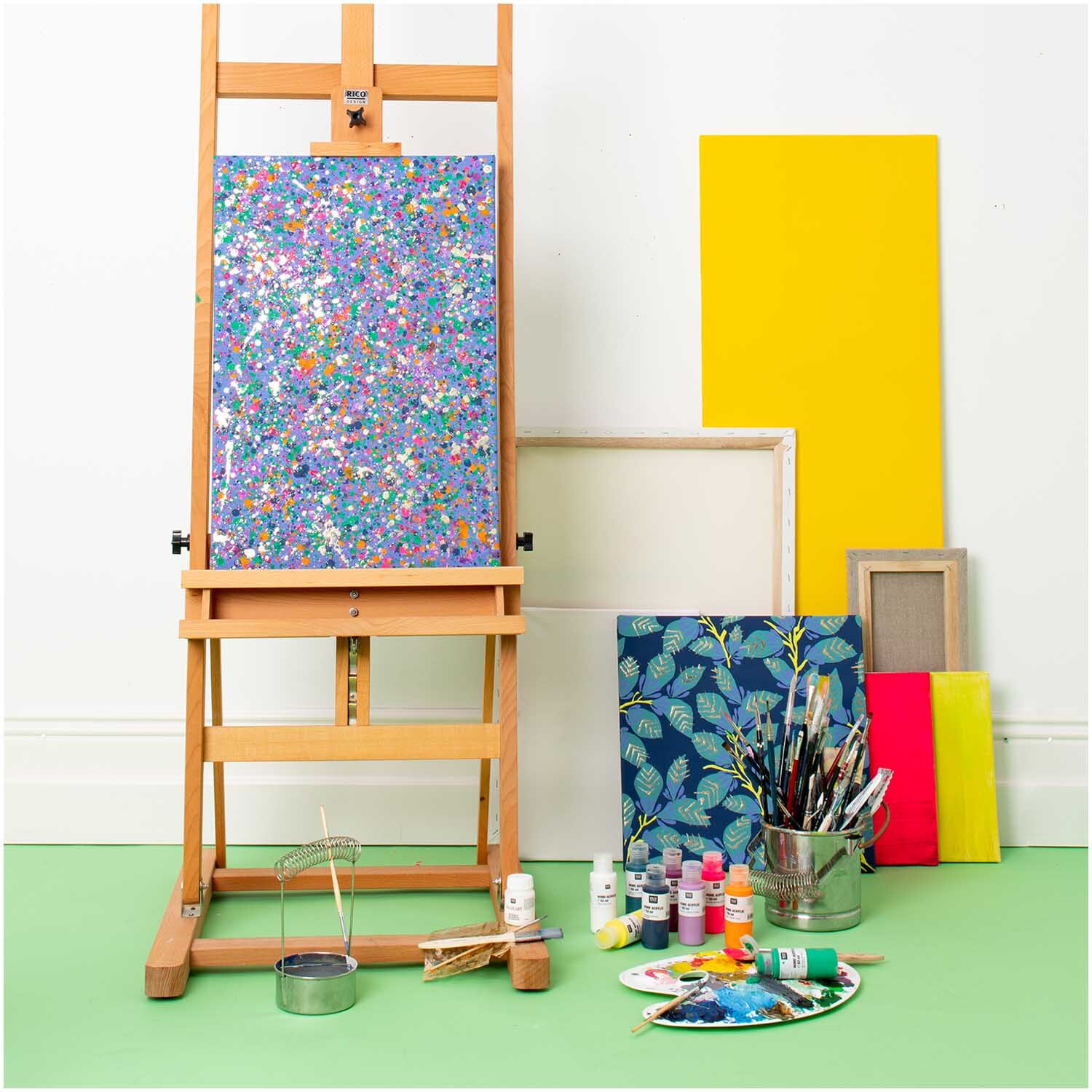 Home Acrylic: Bunte Malutensilien auf grünem Boden, Staffelei mit farbenfrohem Bild, gelbe Leinwand.