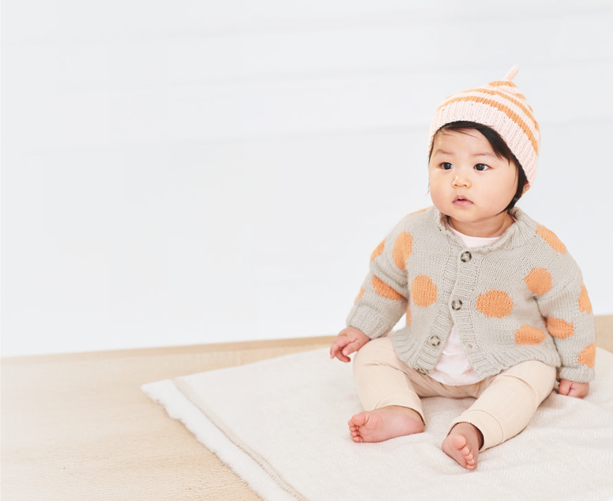 Ein Baby in gestrickter Jacke mit Punkten und gestreifter Mütze sitzt auf einer Decke. Cover des Handstrickmagazins Rico Baby 042 daneben