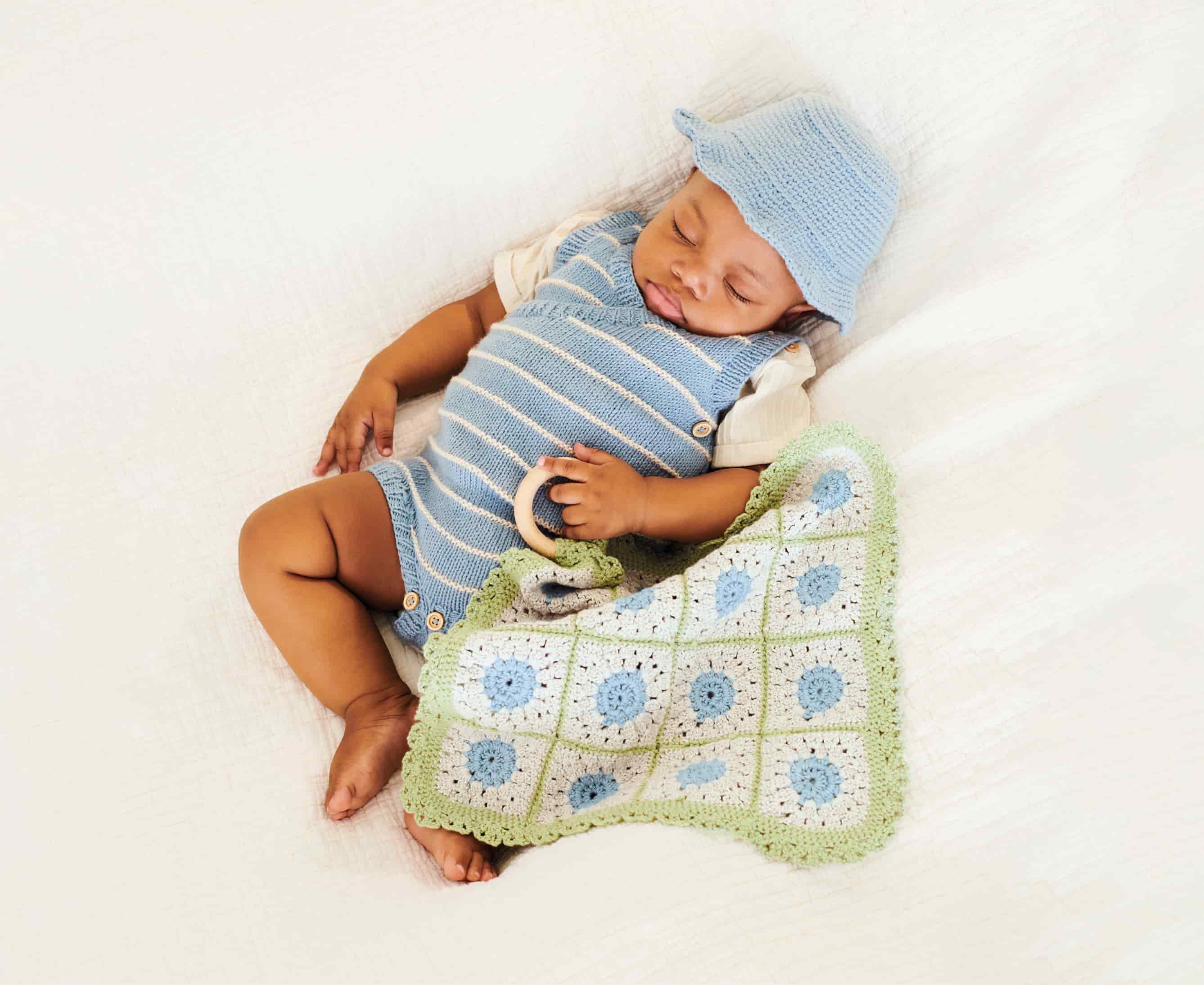 Baby mit gestricktem Outfit und Mütze hält ein gehäkeltes Tuch in der Hand und liegt auf einer Decke.