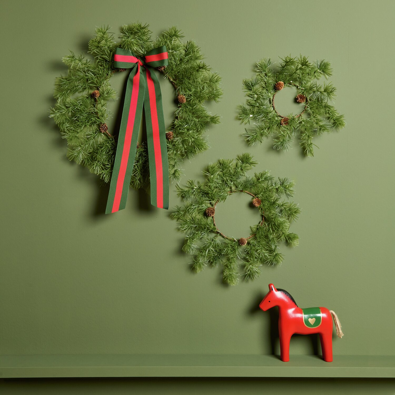 Lärchenkranz mit Zapfen, dekoriert mit roten und grünen Bändern, an grüner Wand.