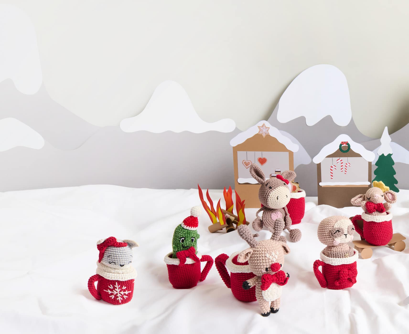 Weihnachlich geschmückte Amigurumis mit gehäkelten Tassen sind in einer aus Papier gestalteten Winterlandschaft dekoriert. 