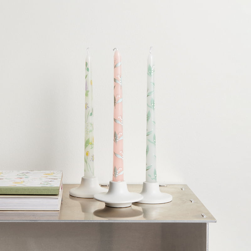 Drei Stabkerzen in Pastelltönen mit Blumenmotiven stehen in Kerzenhaltern dekoriert auf einem Tisch.