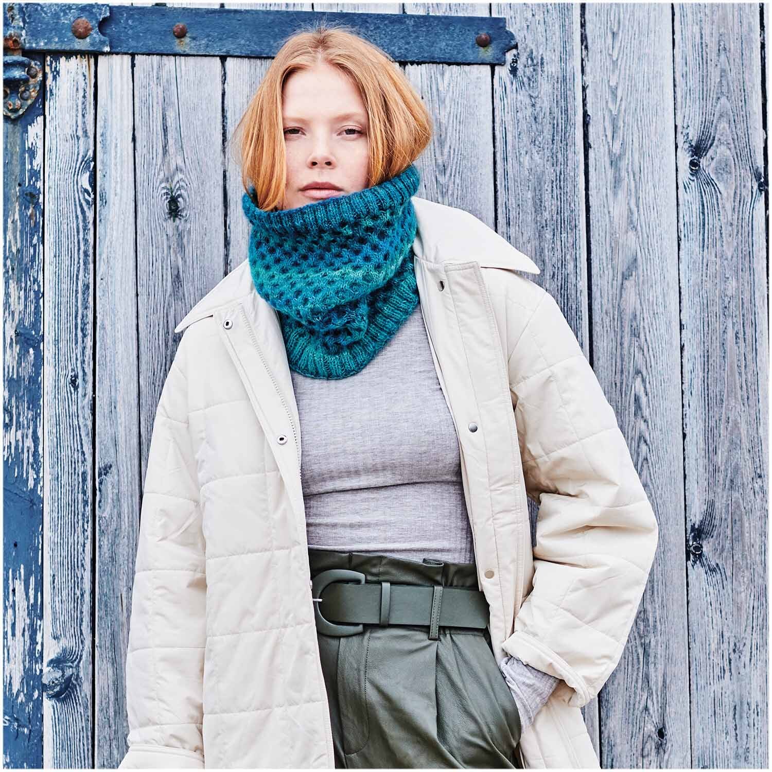 Creative Melange Garzato aran Wonderball: Frau trägt blauen Strickschal vor Holzwand, modisch kombiniert mit beigem Mantel und grauem Oberteil.