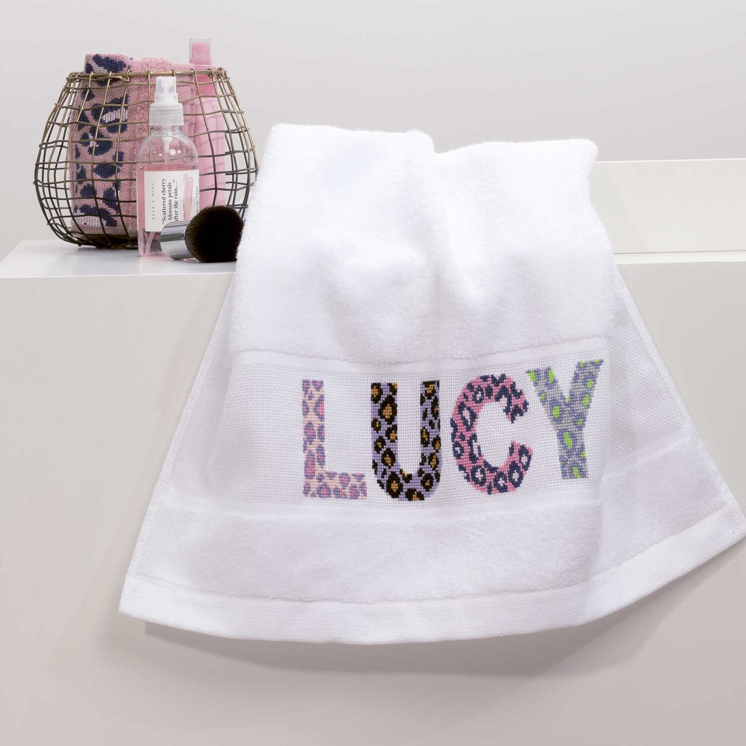 Handtuch 50x100cm Farbe Weiß von Rico Design Handtuch 50x100cm mit buntem "LUCY"-Schriftzug, dekorativ auf einem Regal neben Pflegeprodukten platziert.