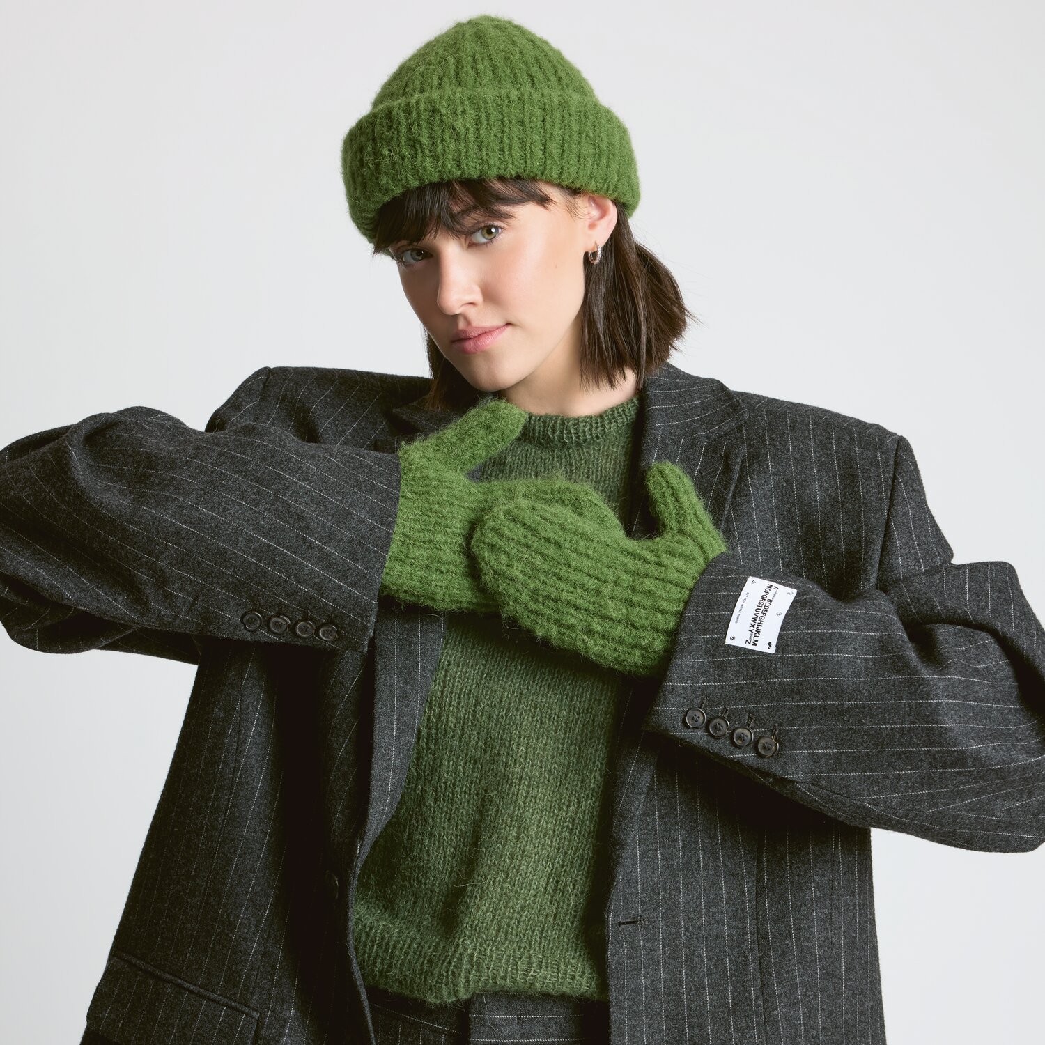 Strickset Mütze Modell 19 aus Luxury Knits - Premium Natural Fibres von Rico Design Strickset Mütze Modell 19 aus Luxury Knits: Frau trägt grüne Mütze und Handschuhe, grauer Anzug.