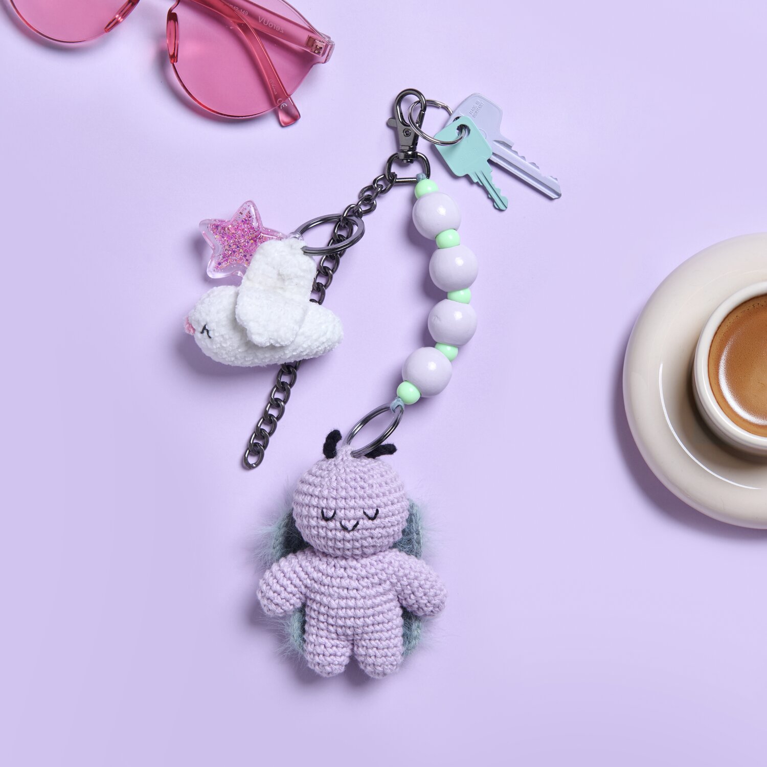 Karabiner mit Kette und Schlüsselring 13cm von Rico Design Karabiner mit Kette und Schlüsselring 13cm, silber, mit gehäkeltem Anhänger und Perlen, auf lila Hintergrund neben Sonnenbrille und Kaffeetasse.