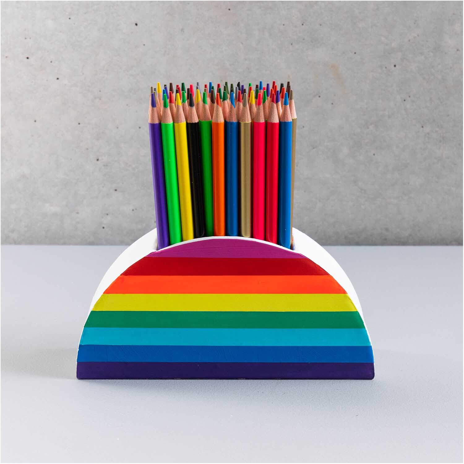 Stiftehalter aus Pappe in halbrunder Form mit Regenbogenstreifen, gefüllt mit bunten Stiften.