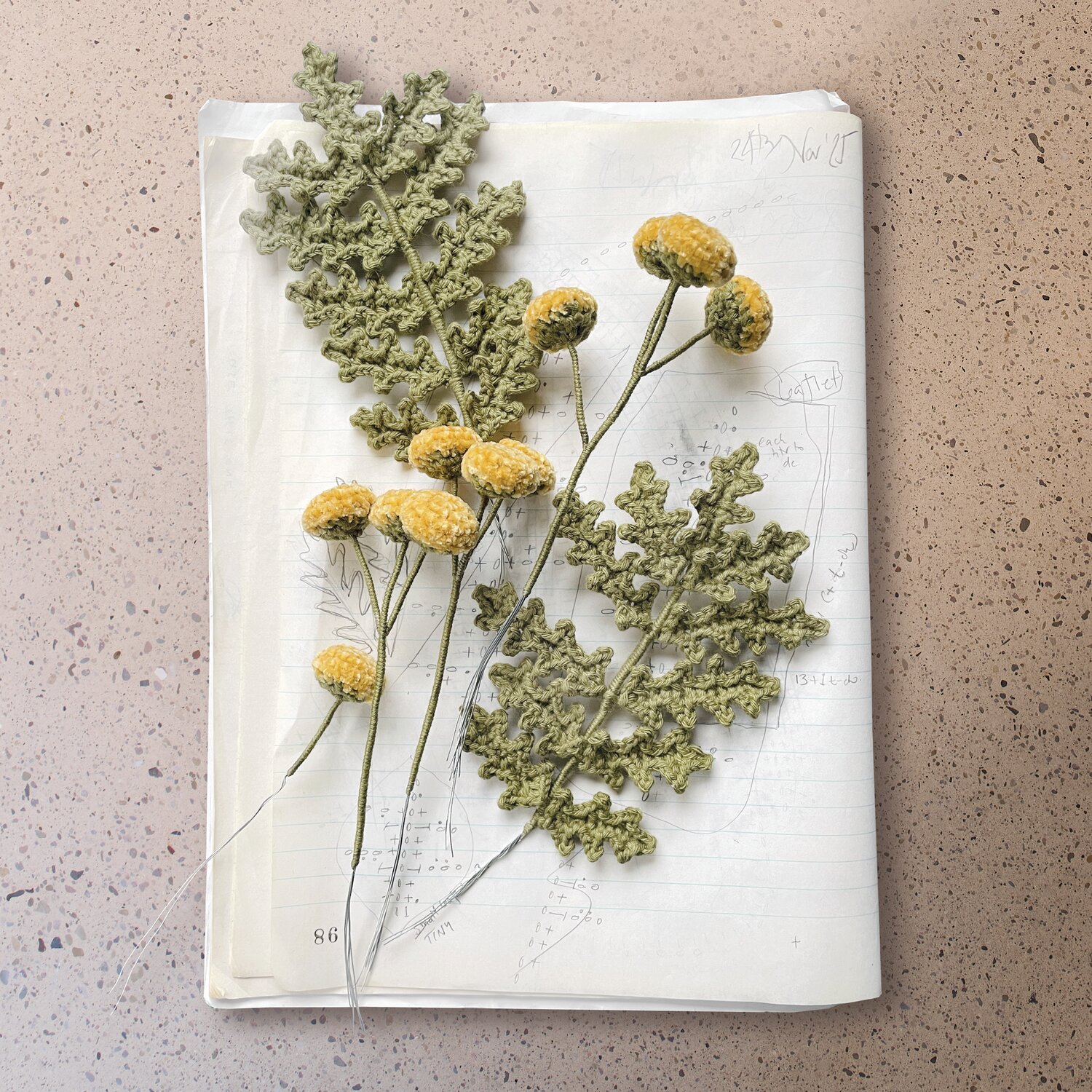 Ricorumi In the Meadow zeigt detailreich gehäkelte Blumen auf aufgeklapptem Skizzenbuch, dekorativ arrangiert zur Inspiration für kreative Häkelprojekte