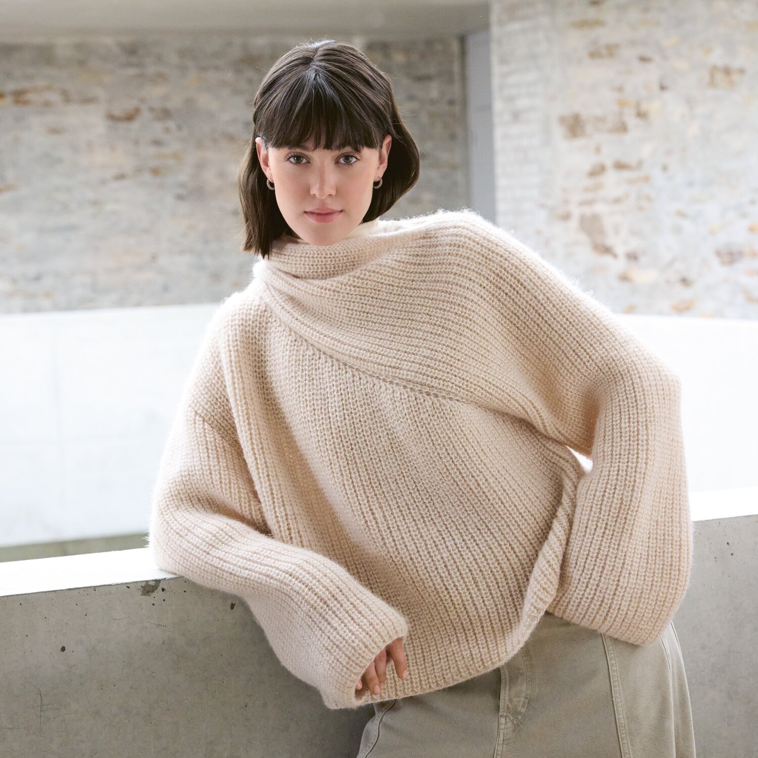 Strickset Pullover Modell 01 aus Luxury Knits - Premium Natural Fibres von Rico Design Strickset Pullover Modell 01 aus Luxury Knits zeigt eine Frau in einem cremefarbenen Pullover vor einer Steinwand.