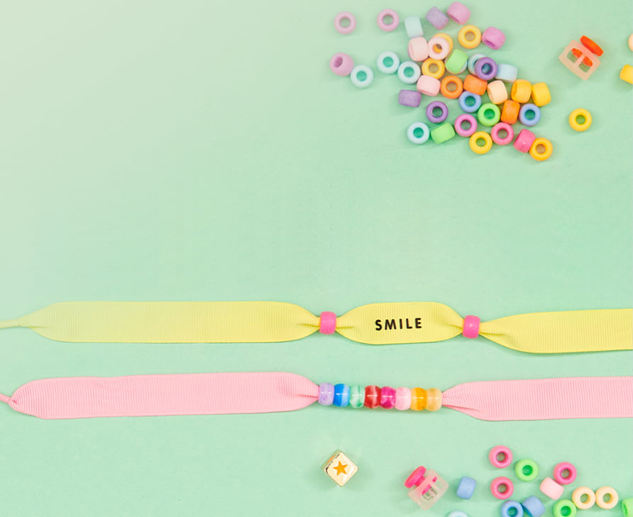 Zwei Ponii Beads Ripsbänder in Gelb und Rosa sind mit bunten Großlochperlen geschmückt und liegen auf hellgrünem Hintergrund. Darum herum liegen weitere bunte Perlen