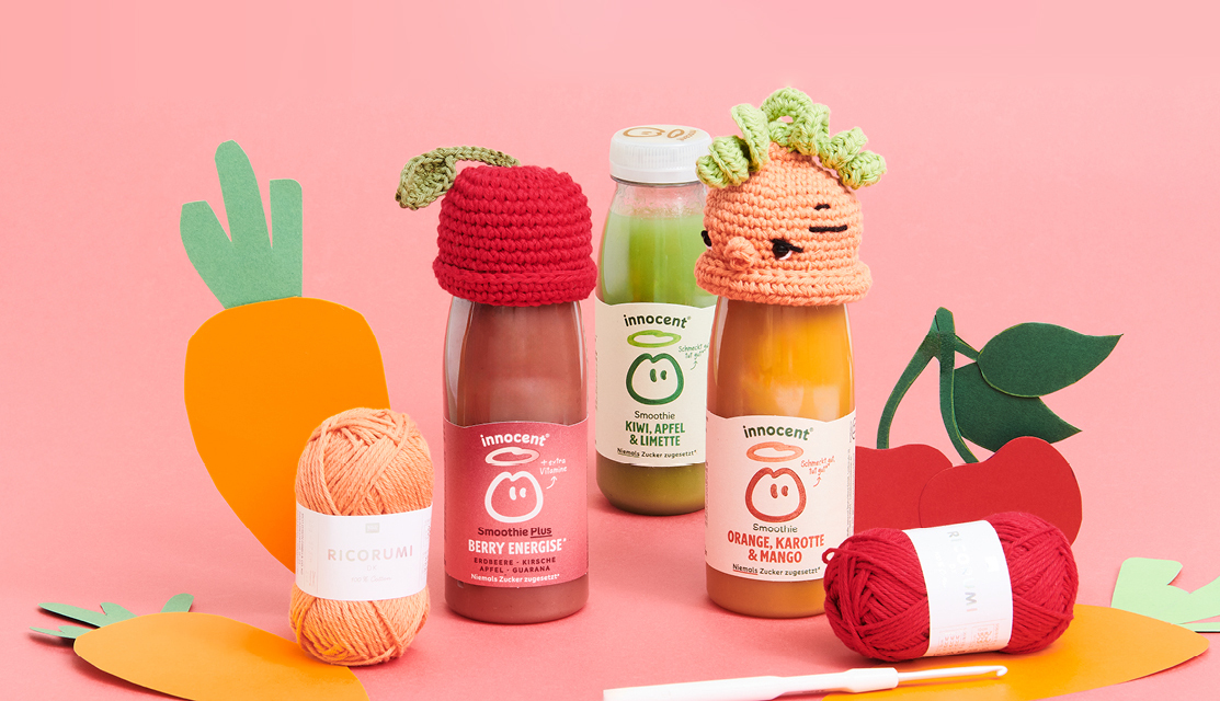 Häkelanleitung Ricorumi Mützchen Kirsche: Bunte Smoothie-Flaschen mit gehäkelten Mützen auf rosa Hintergrund.