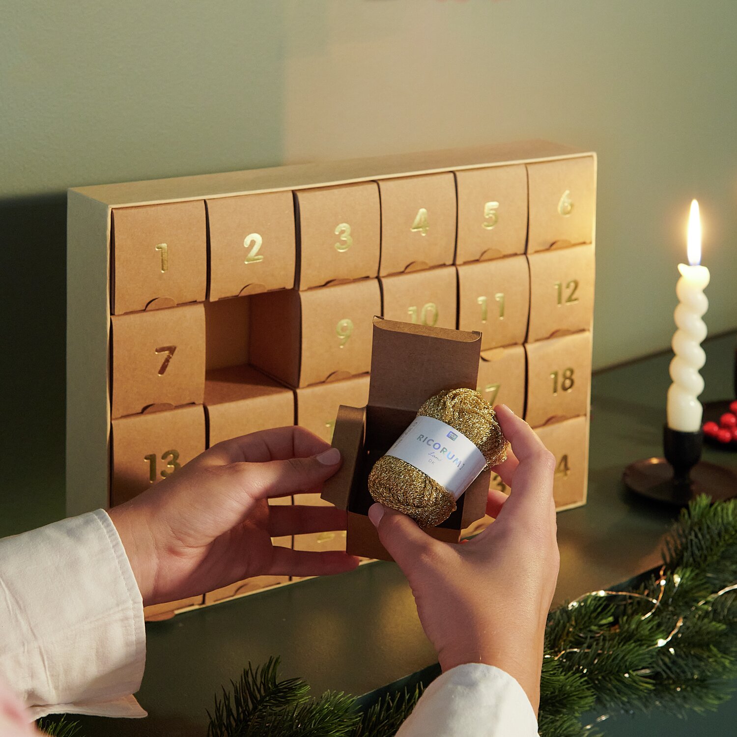 Ricorumi Adventskalender: Person öffnet braune Box mit goldenem Garn von Rico Design, Kerze im Hintergrund.
