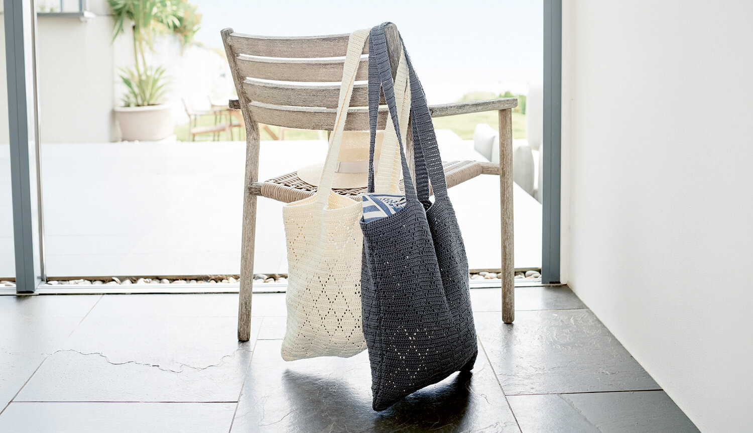 Häkelanleitung Lochmuster-Tasche aus Essentials Organic Cotton aran zeigt zwei gehäkelte Schultertaschen mit feinem Lochmuster, die dekorativ an einem Holzstuhl über einem hellen Fliesenboden hängen.