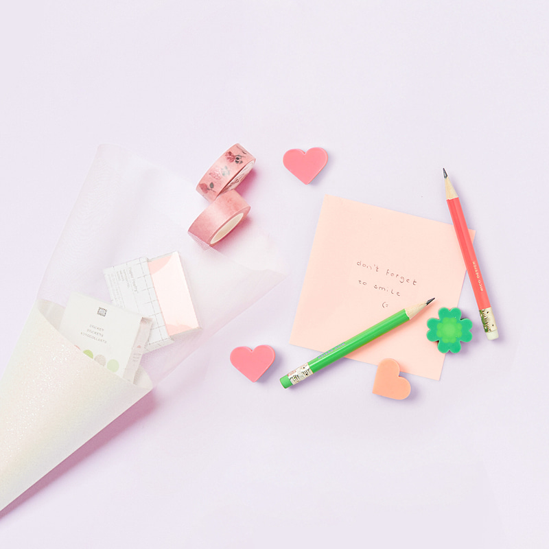 Schultütenrohling mit weißem Glitter liegt geöffnet und umgeben von Tapes, Post Its, Radiergummis und Stiften auf lila Hintergrund