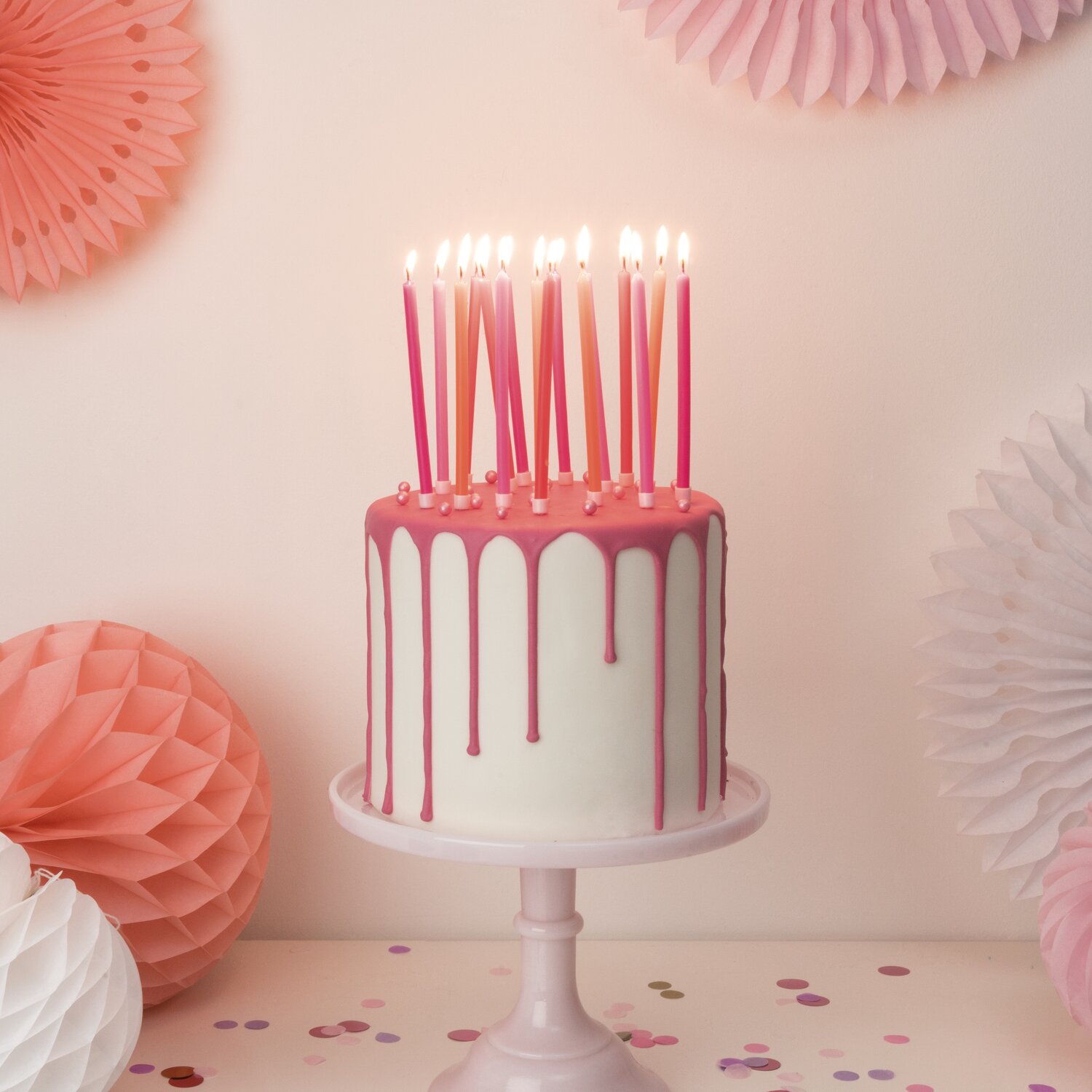 Kerzen lang 12cm, Rico Design, 16 Stück, auf weiß-rosa Drip-Torte mit brennenden Neonstäben, dekoriert mit Wabebällen und Konfetti auf hellem Tisch