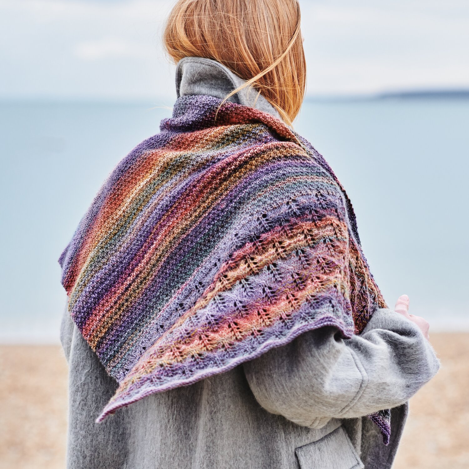 Strickset Tuch Modell 08 aus Wonderball Special: Frau am Strand mit buntem, gestricktem Tuch in Lila- und Brauntönen über grauem Mantel.