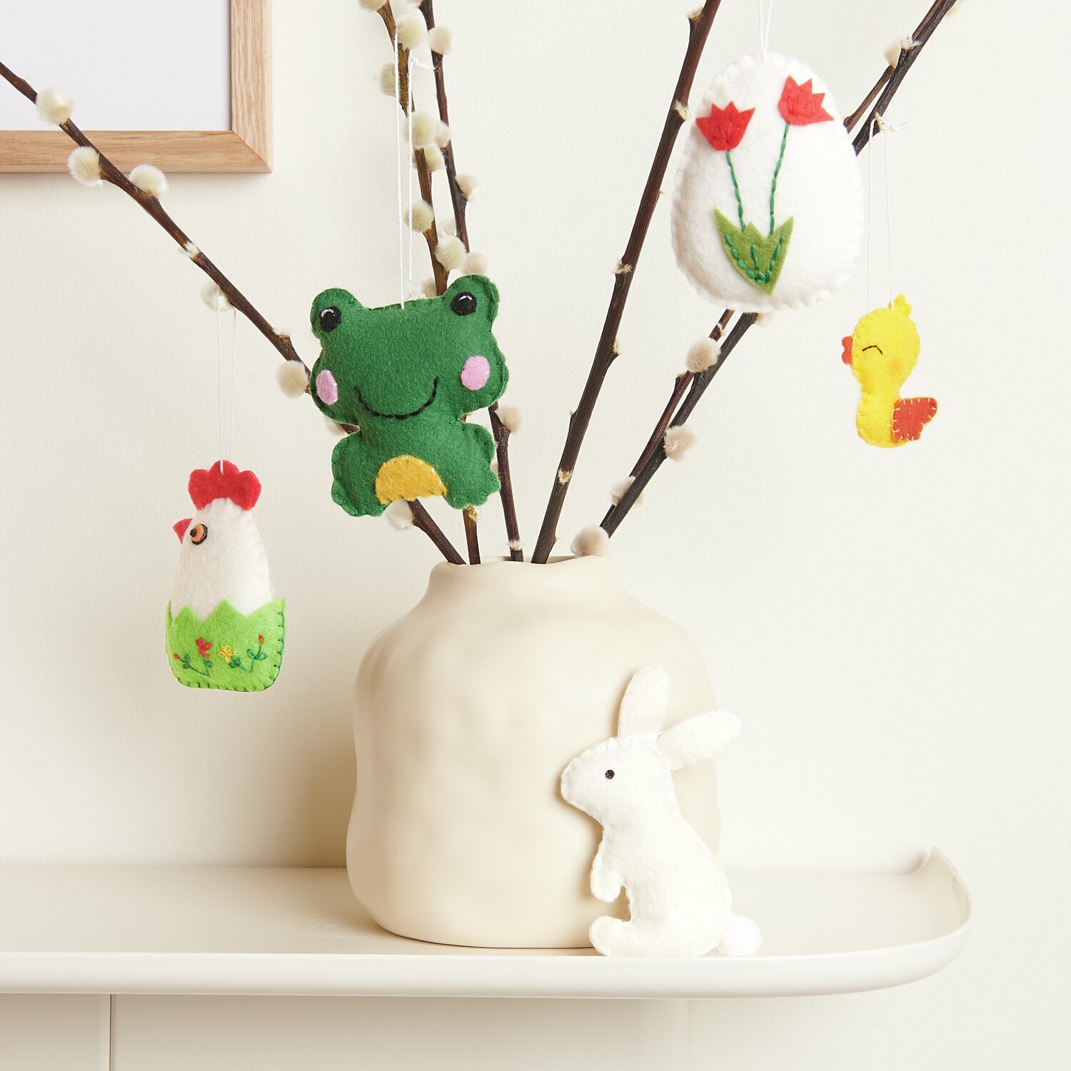 Aufhänger Filz-Osterfiguren 2D 5 Stück: Bunte Filzfiguren an Zweigen in einer cremefarbenen Vase.