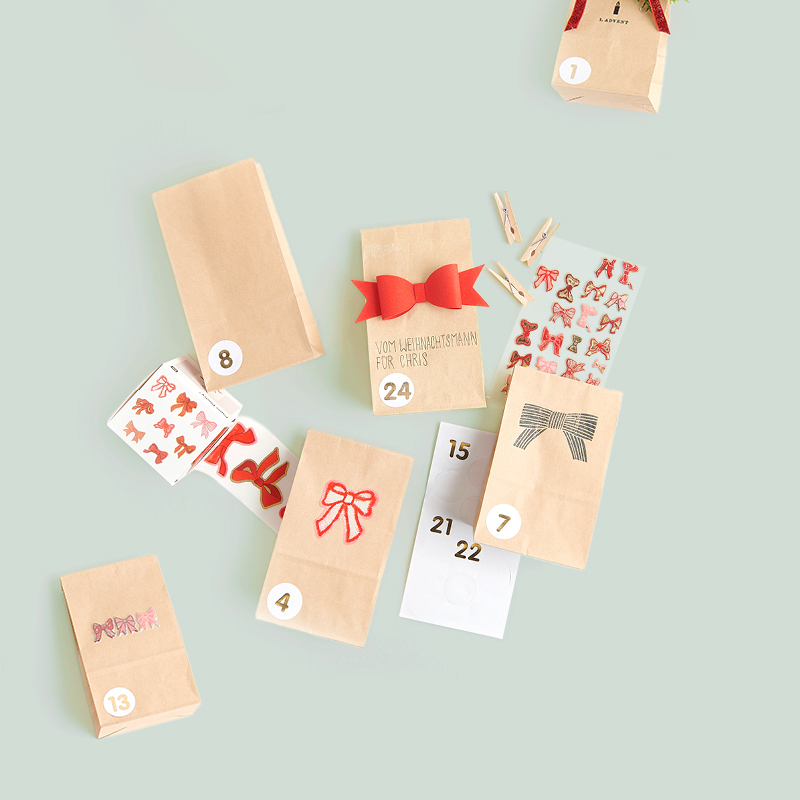 Adventskalender aus Papiertüten, geschmückt mit Zahlen und Stickern liegen ausgebreitet auf grünem Hintergrund