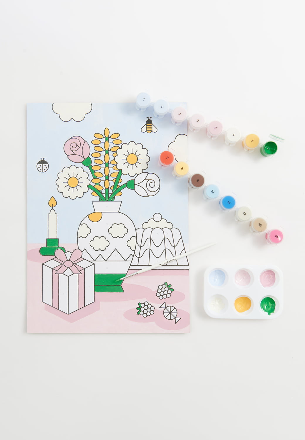 Ein Malen-nach-Zahlen Bild mit einer illustrierten Blumenvase, Kuchen und Geschenk liegt umgeben von Farbtöpfen auf grauem Hintergrund. 