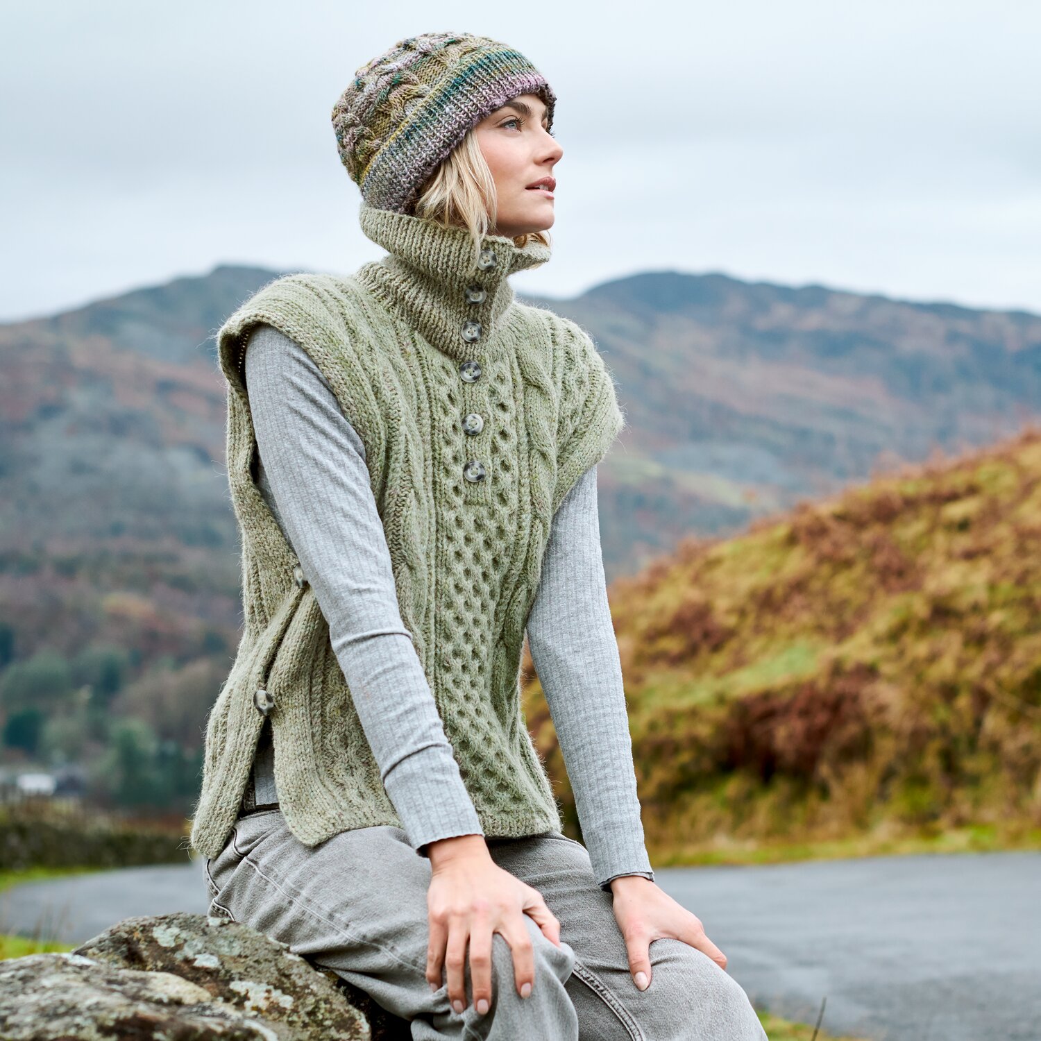 Strickset Pullunder Modell 09 aus Lovewool No. 23, ärmelloser Zopfpullunder mit Stehkragen, seitlichen Knöpfen und passender Mütze, getragen in herbstlicher Berglandschaft