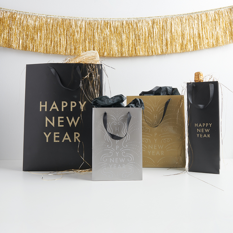 Geschenktüten in Schwarz, Silber und Gold und Schriftzug "Happy New Year" nebeneinander aufgestellt vor goldener Girlande