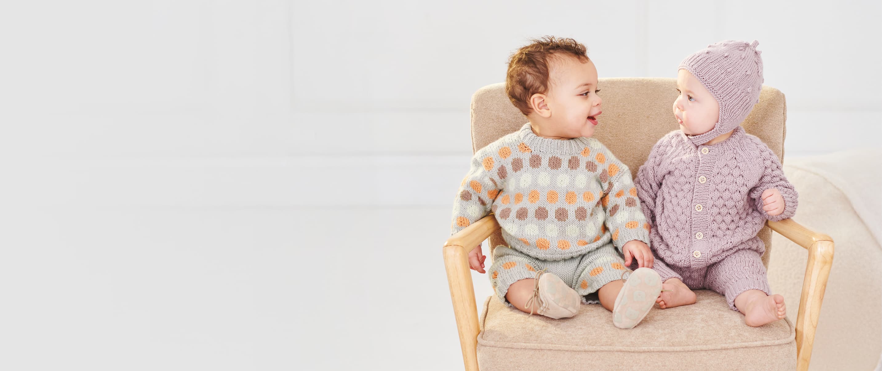 Zwei Babys sitzen in Strickkleidung gekleidet auf einem Stuhl und schauen sich fröhlich an
