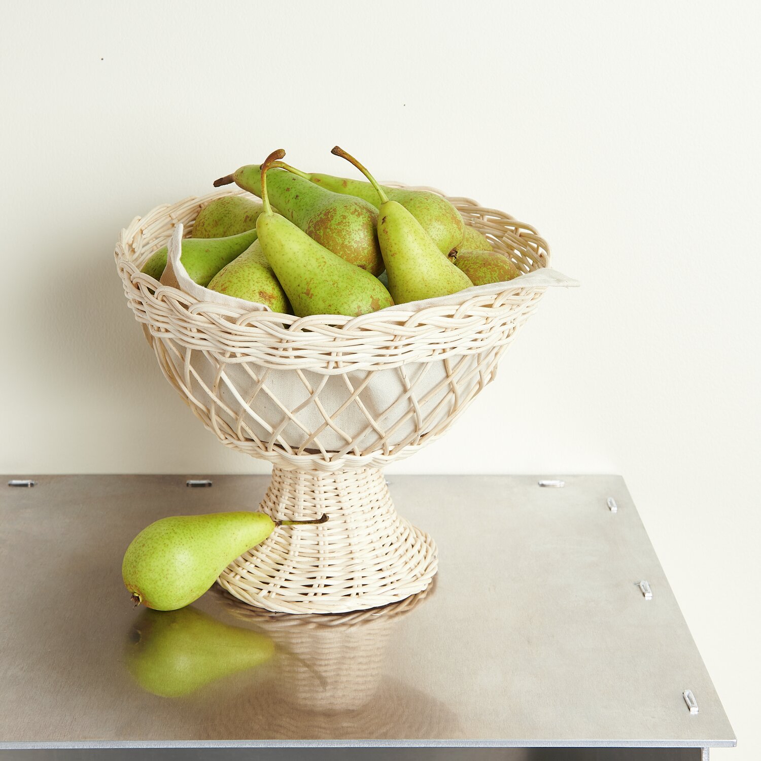 Rattan Korb mit Fuß Weiß, Ø26x22cm, gefüllt mit grünen Birnen auf einem Metalltisch.