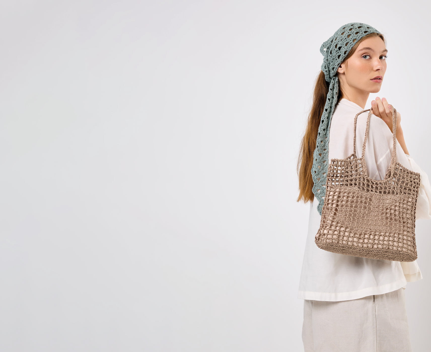 Eine Frau trägt ein sommerliches Outfit mit gehäkeltem Tuch und gehäkelter Tasche und posiert vor grauem Hintergrund.