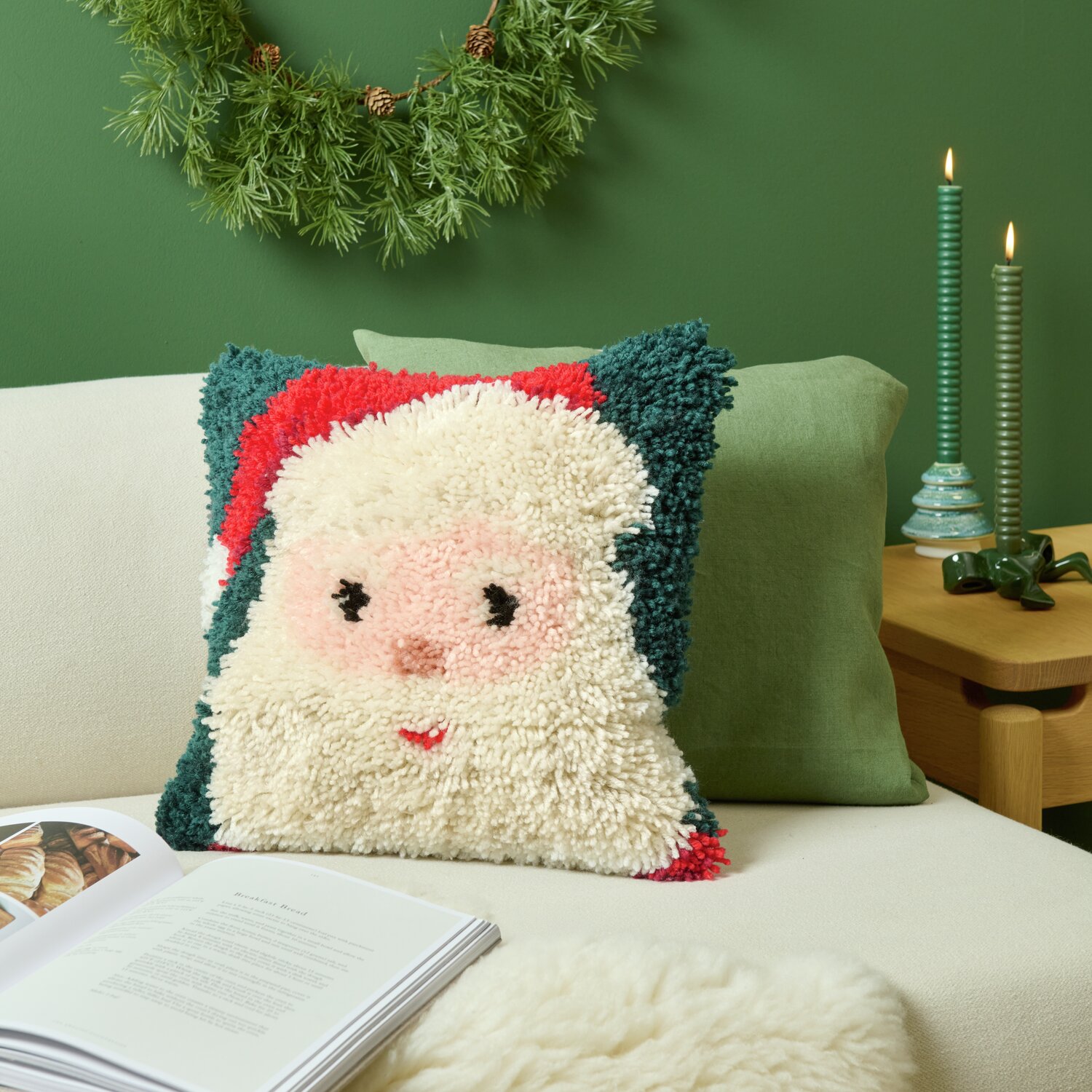 Rico Hooki Hooki Knüpfset Kissen Santa zeigt ein weihnachtliches Design mit Santa-Motiv auf einem Sofa, umgeben von Dekoration.