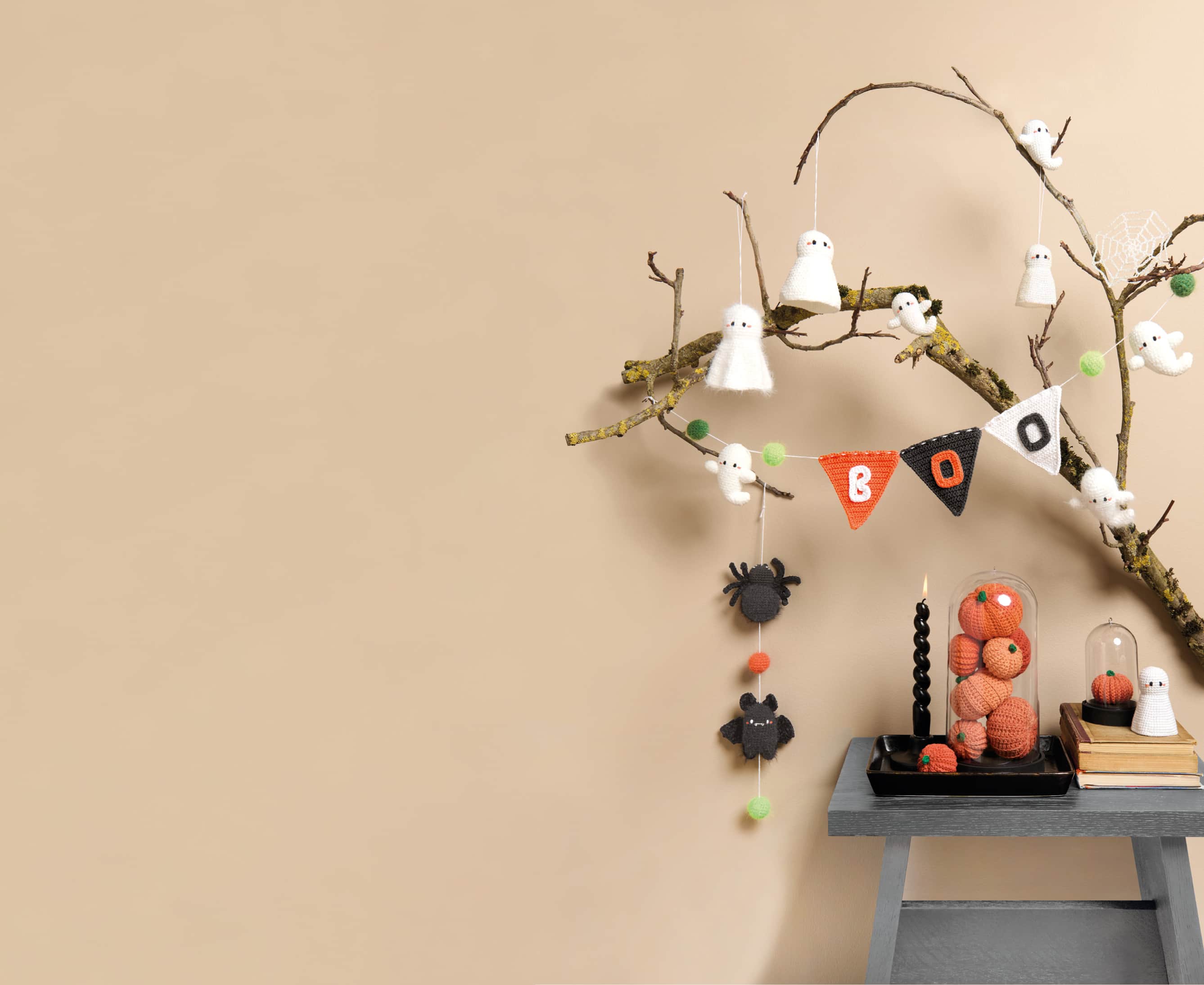 Gehäkelte Halloweenfiguren in Form von Kürbissen, Gespenstern und Spinnen sind auf einem Tischchen und an einem Ast dekoriert. 