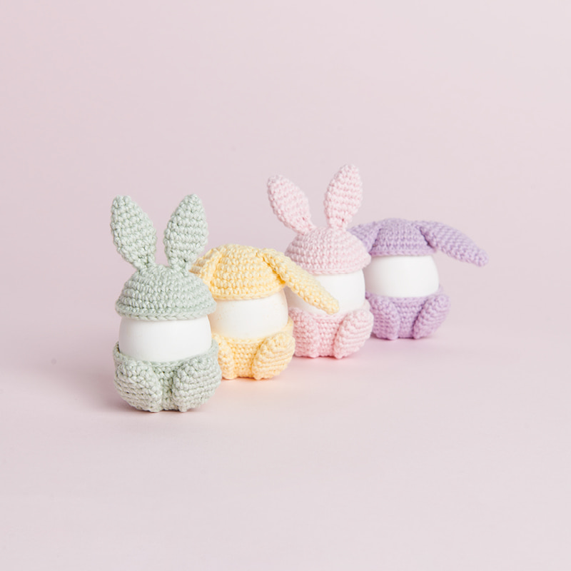 Vier pastellfarbene, gehäkelte Eierwärmer in Hasenform stehen auf rosa Hintergrund.