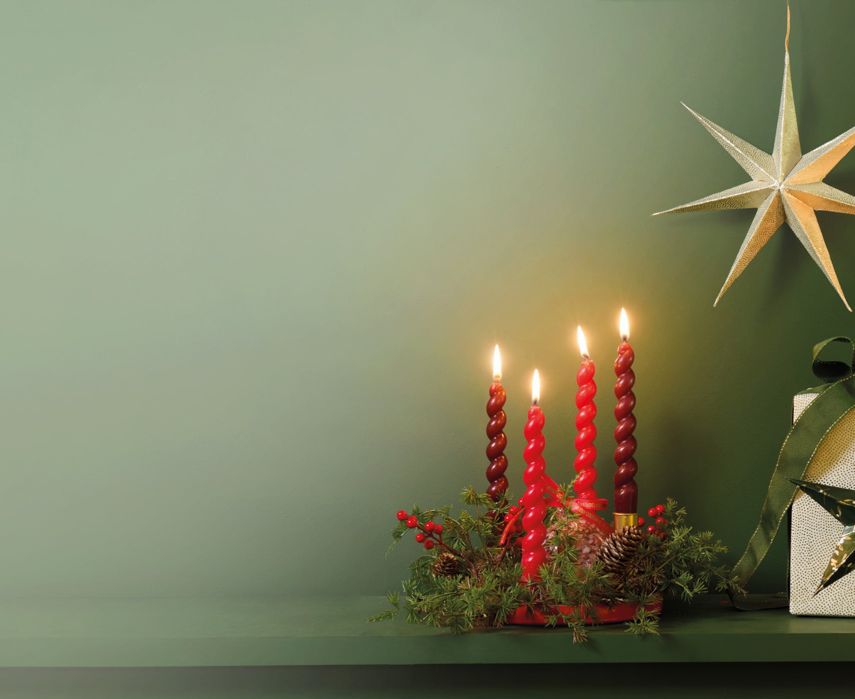 Moderner Adventskranz mit vier roten Kerzen, Tannengrün und roten Beeren vor grüner Wand. Daneben stehen ein Geschenk und ein leuchtender Papierstern.