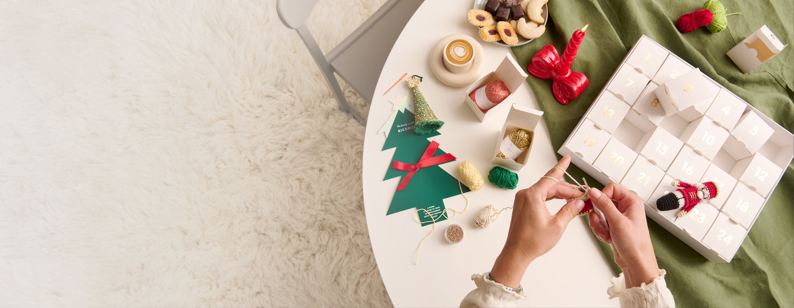 Der Ricorumi Adventskalender liegt geöffnet auf einem Tisch. Einzelne Adventskalender-Boxen liegen geöffnet daneben. Hände häkeln an einem Häkelmodell