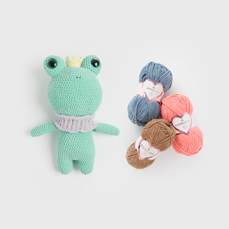 Ein gehäkelter Frosch liegt neben drei Garnknäueln von Creative Chenillove auf grauem Hintergrund.