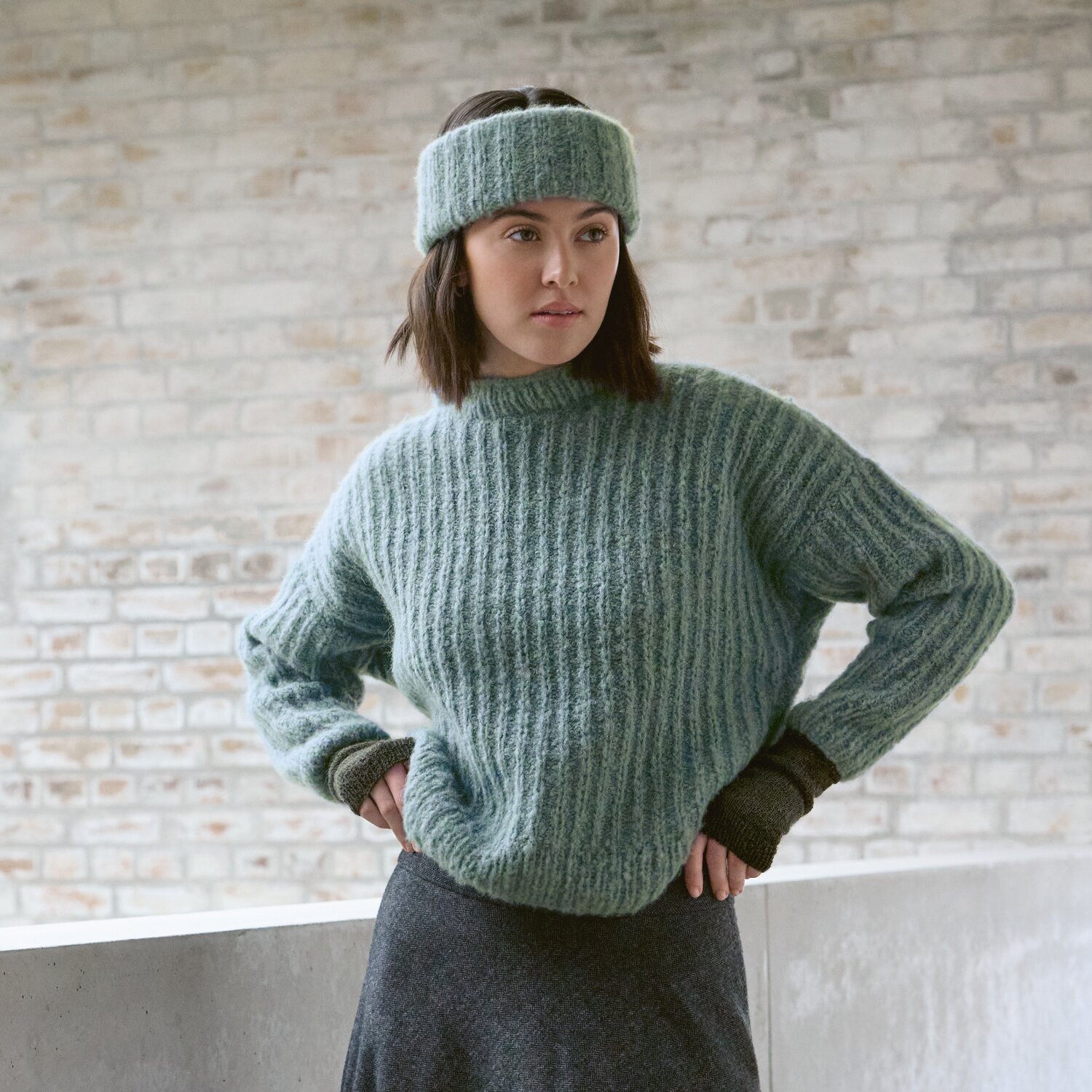 Strickset Pullover Modell 12 aus Luxury Knits - Premium Natural Fibres von Rico Design Strickset Pullover Modell 12 aus Luxury Knits: Frau trägt grünen Pullover und Stirnband vor Backsteinwand.