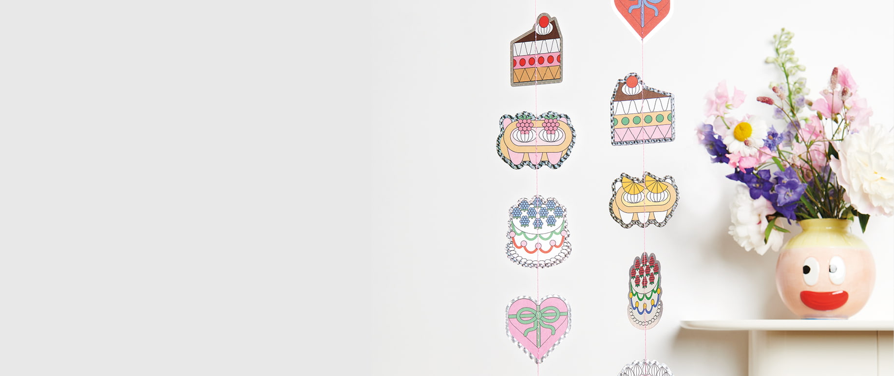 Eine Girlande mit Motiven von Torten und Kuchen hängt an einer Wand neben einer dekorierten Blumenvase.