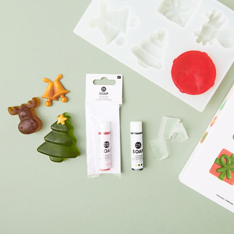 Seifen in weihnachtlichen Formen liegen zusammen mit Rohseife, Silikonformen und Seifenfarbe auf hellgrünem Hintergrund.