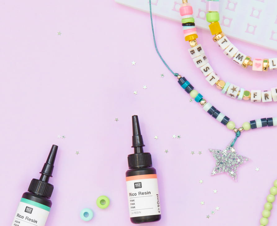 Perlen und Schmuckstücke aus Resin sind auf Ketten aufgefädelt. Flaschen von UV Resin und eine Silikonform liegen daneben auf rosa Hintergrund