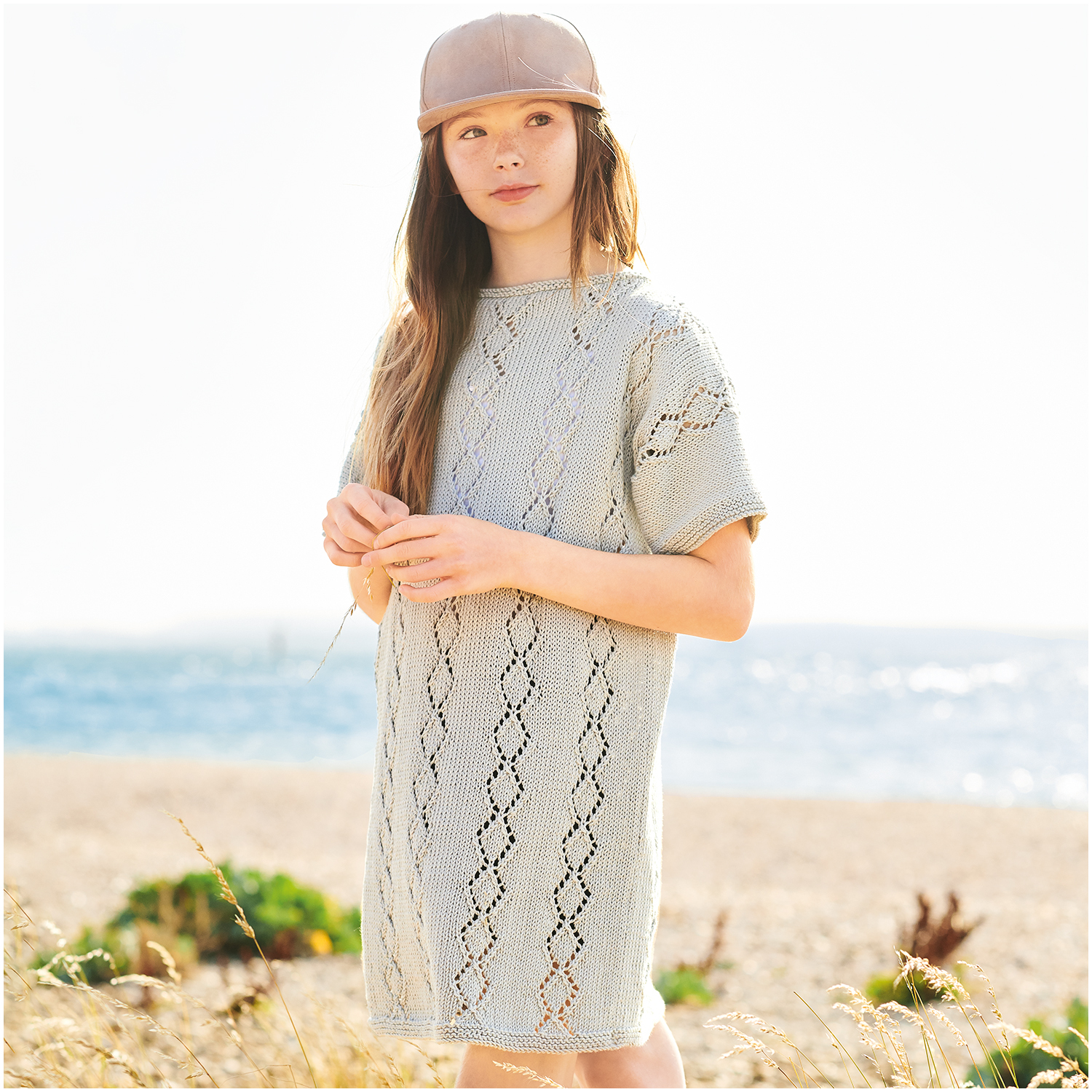 Essentials Organic Cotton dk: Mädchen in gestricktem Kleid am Strand, trägt eine Kappe, im Hintergrund Meer.