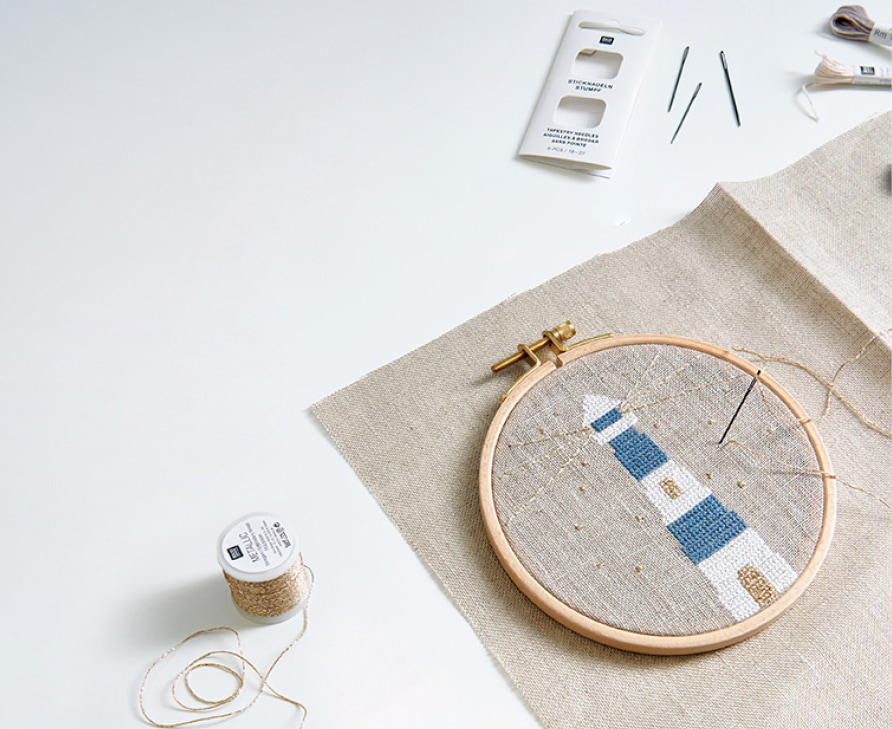  Ein auf Stoff gestickter Leuchtturm in weiß, blau und gold ist in einen Stickrahmen gespannt und umgeben von weiterem Stickzubehör