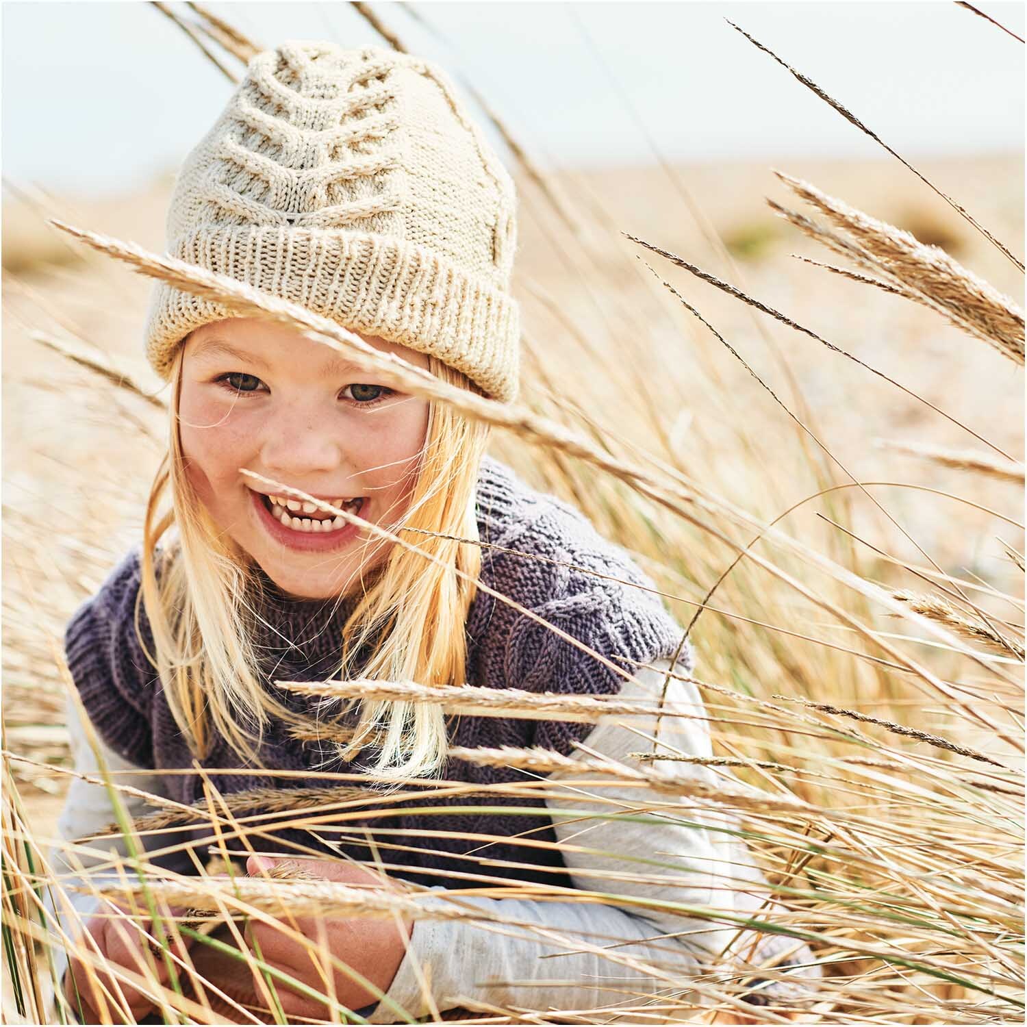 Essentials Merino dk: Ein lächelndes Kind trägt eine gestrickte Mütze in einem Feld aus hohem Gras.
