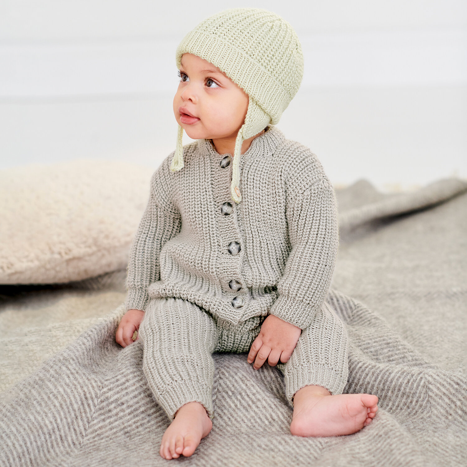 Strickanleitung Mütze und Strampler aus Rico Baby Dream dk uni, sitzendes Baby im gestrickten Einteiler mit passender Mütze auf weicher Decke in hellen Naturtönen