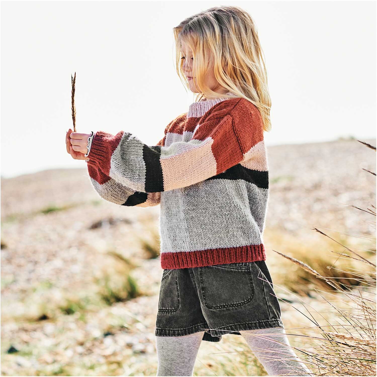 Essentials Super aran 50g 100m Farbe Rost von Rico Design Essentials Super aran: Kind in gestreiftem Pullover aus Rico Design Wolle, spielt draußen an einem sonnigen Tag.