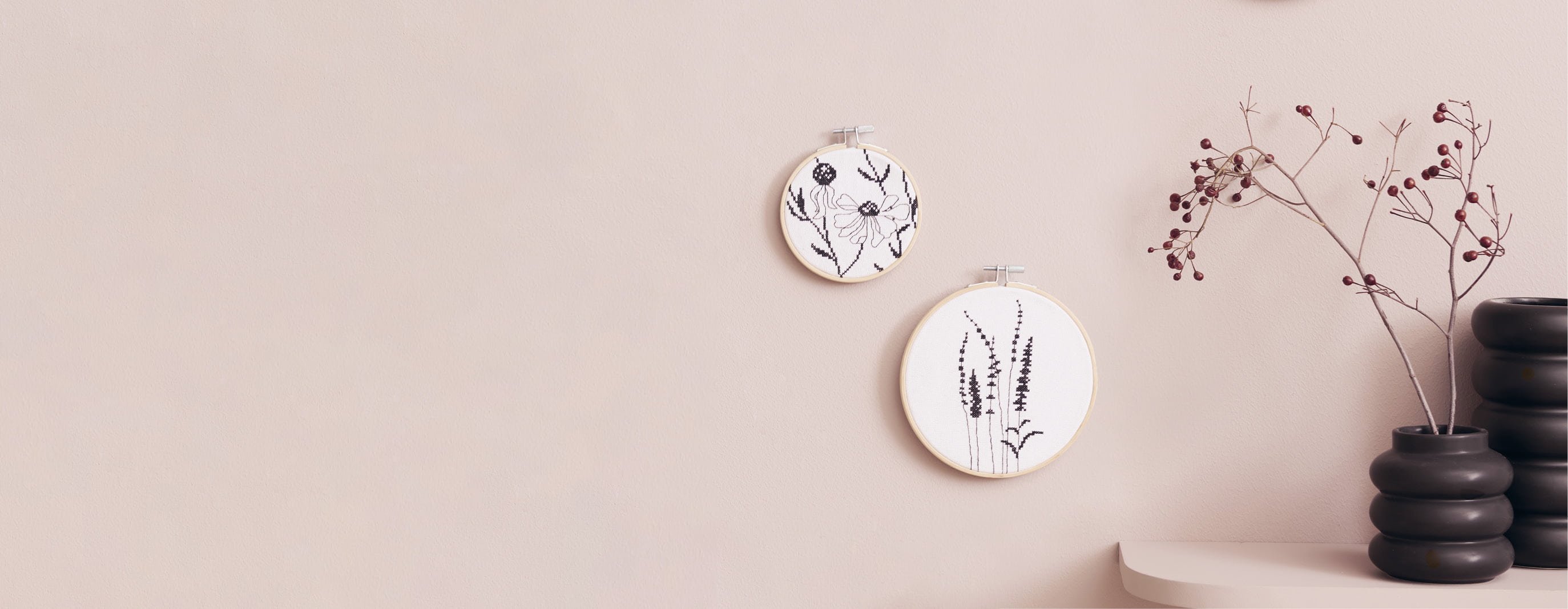 Stickbilder mit Gräser- und Blumenmotiven hängen an einer beigen Wand. 