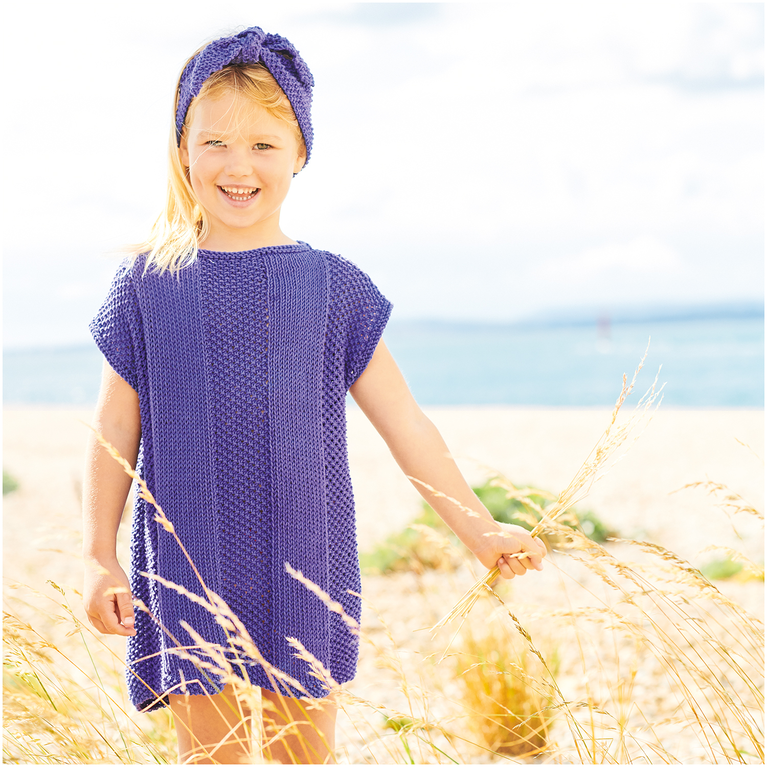 Essentials Organic Cotton aran: Mädchen in violettem Strickkleid und Stirnband am Strand, Rico Design.