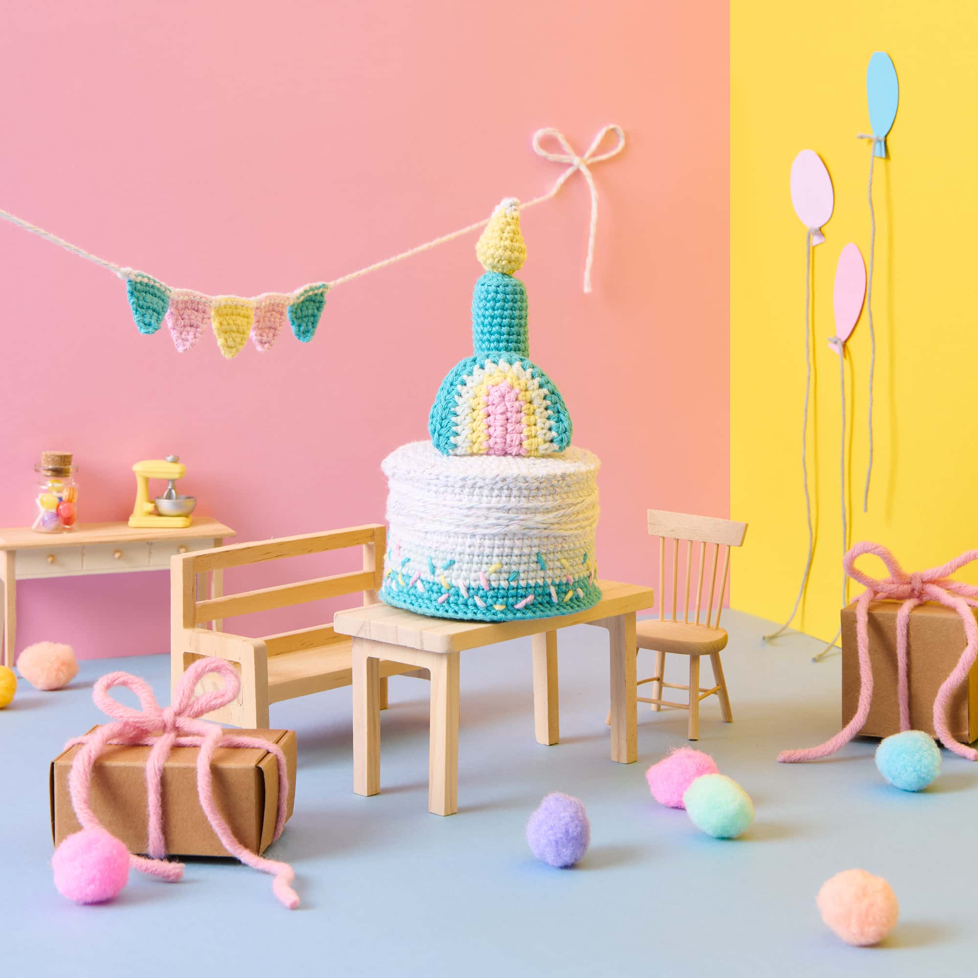 Eine gehäkelte Torte mit Regenbogen und Kerze steht auf einem Tisch in einer bunt dekorierten Szene mit Partydekoration.