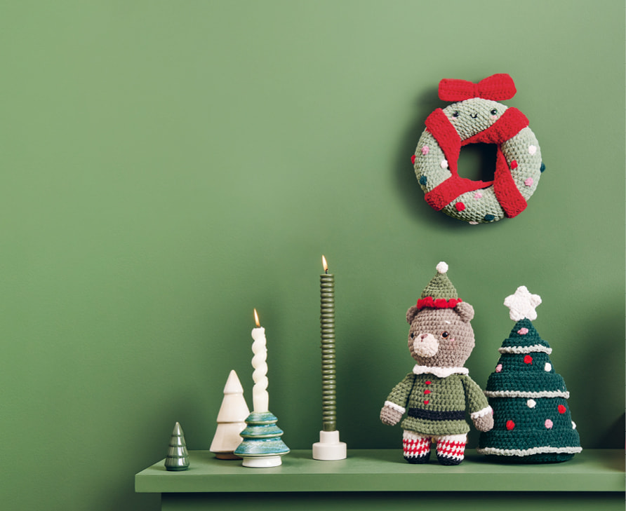 Weihnachtliche Häkelfiguren aus Chenille-Garn sind dekoriert auf einem Regal und an einer grünen Wand dekoriert