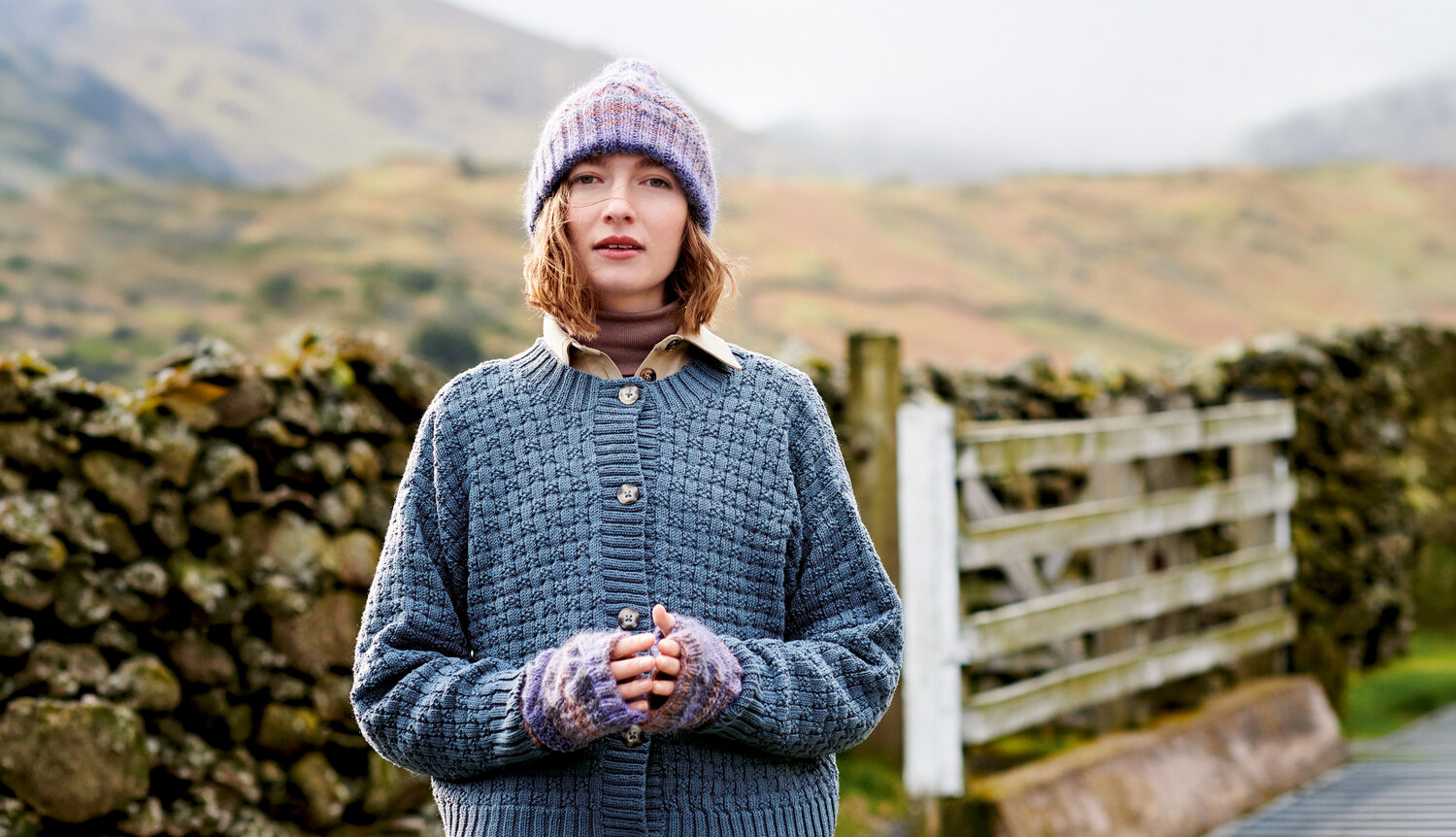 Strickanleitung Mütze und Stulpen aus Creative Alpaca Melange aran, getragen von einer Frau im ländlichen Herbstpanorama mit Steinmauer und Holzzaun im Hintergrund